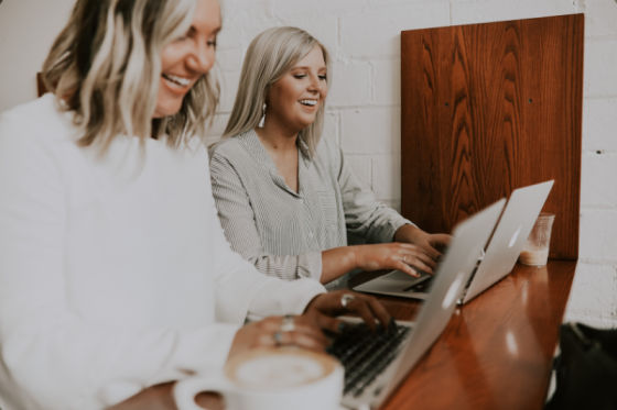 Two women smiling as they work at a counter on their laptops.