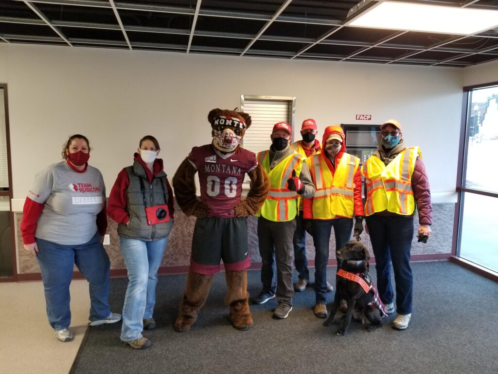 Men and women in COVID face masks pose with a University of Montana bear mascot while volunteering at a COVID vaccine clinic