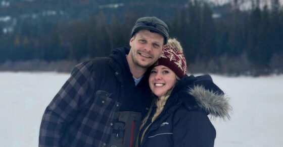 Young man and woman in winter clothes and hats smile in front of a wintry mountain landscape